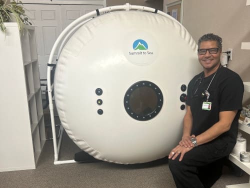 A man sitting beside a large hyperbaric oxygen therapy chamber.